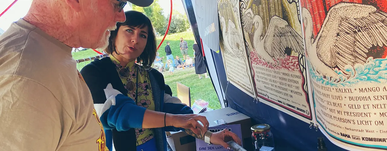 Zwei Personen stehen an einem Stand und diskutieren über etwas, das auf dem Tisch ausgestellt ist. Eine Person zeigt auf einen Gegenstand, während die andere zusieht. Im Hintergrund hängen große Plakate. Die Szene scheint im Freien zu sein.