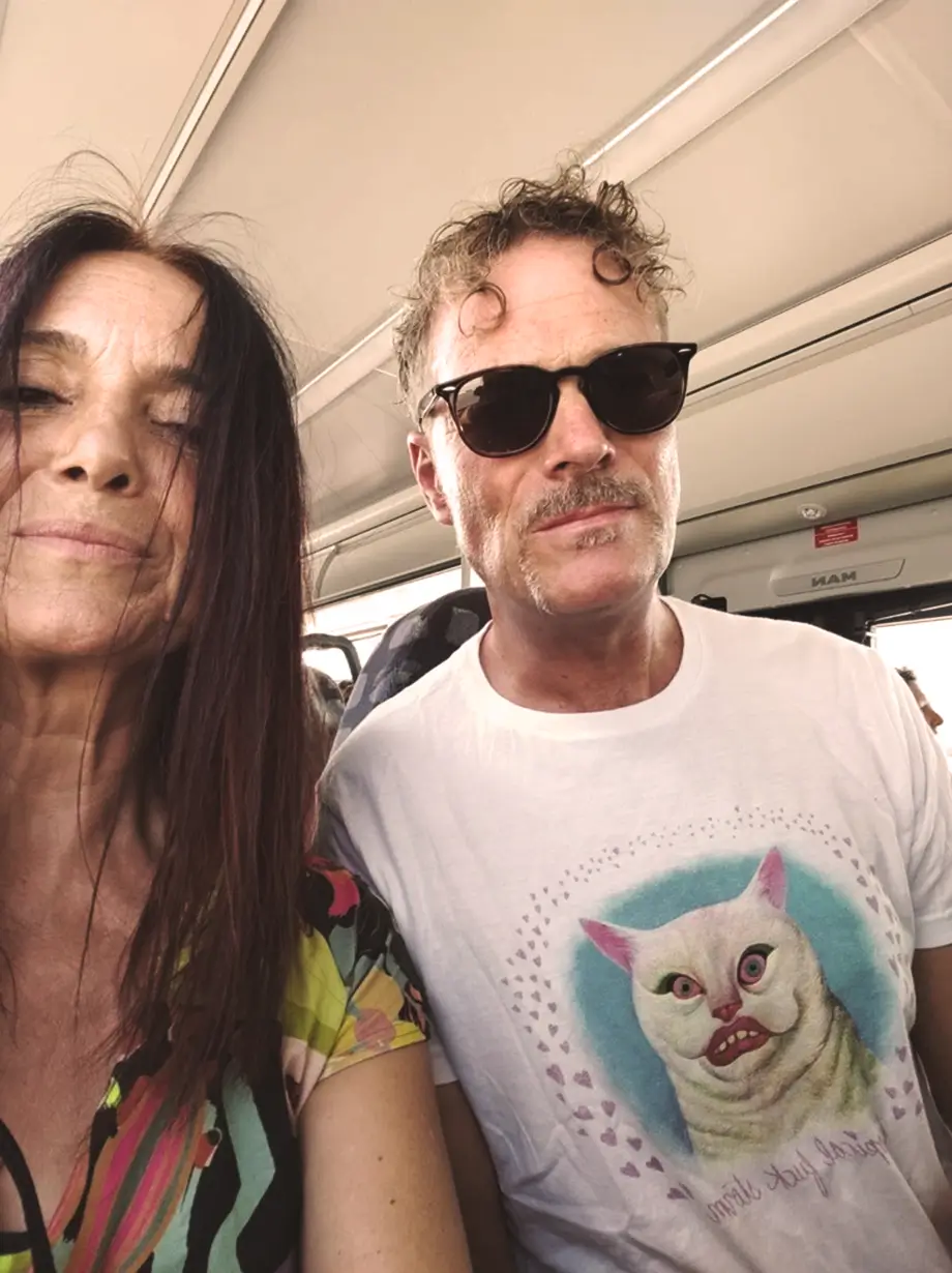 Zwei Erwachsene sitzen nebeneinander in einem Fahrzeug. Der Mann trägt eine Sonnenbrille und ein T-Shirt mit der Abbildung einer Katze mit großen Augen. Die Frau hat langes Haar und trägt ein buntes Oberteil.