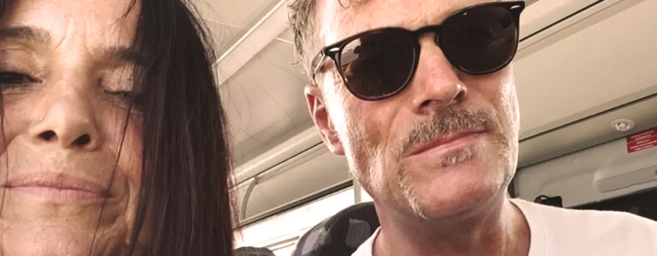 Zwei Menschen sitzen eng beieinander in einem Bus; die Frau hat dunkles Haar, und der Mann trägt eine Sonnenbrille und hat einen Schnurrbart.